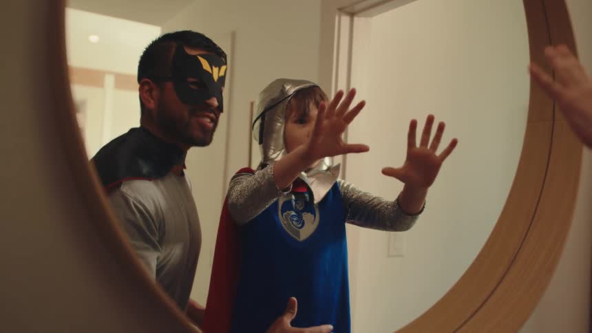A father and son dressed as superheroes strike powerful poses in front of a mirror
