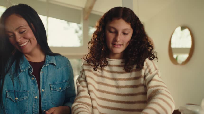 An adult woman and a teenage girl are happy as they are preparing the cookies that are on a table in front of them