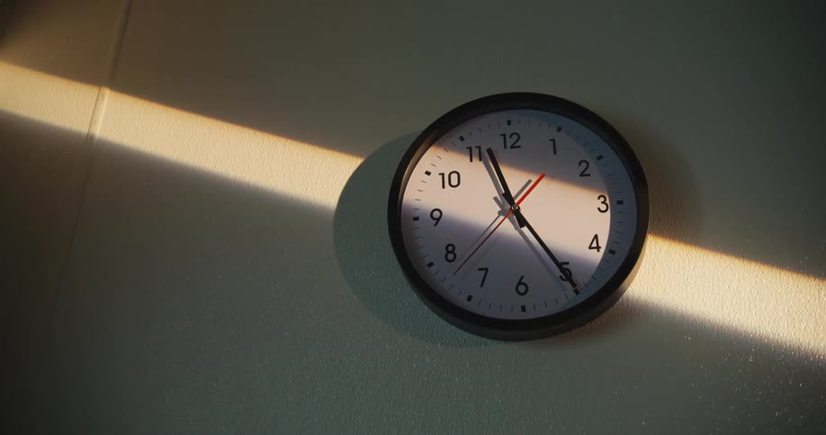 An analog wall clock hangs on a wall with sunlight reflecting off the surface casting a soft glow around it