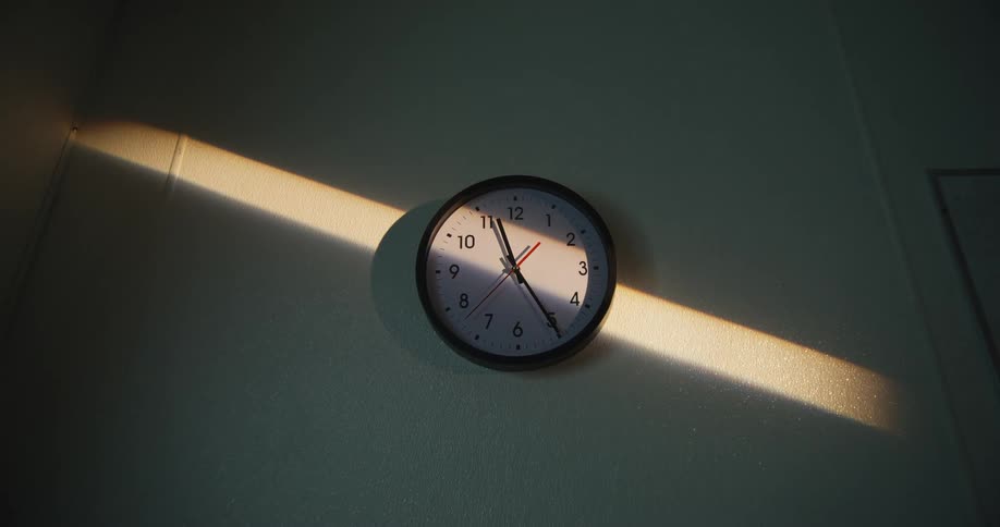 A wall clock is dramatically lit by a diagonal beam of sunlight casting deep shadows in a quiet room