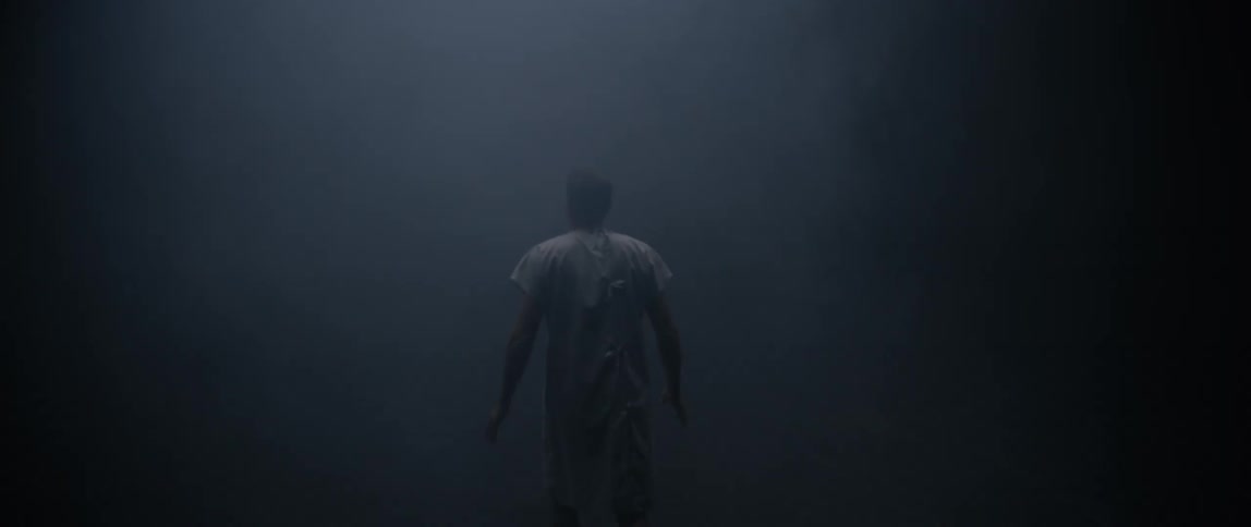 The back of a man wearing a white apron and walking in a room that is completely dark with a light coming from above