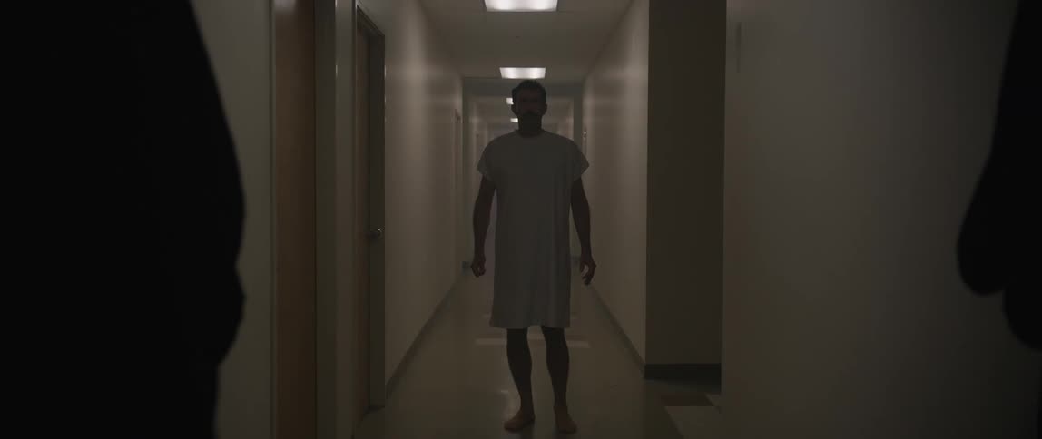 A man in a hospital gown walks into the hallway of a hospital and stands looking forward in a dimly lit hazy corridor framed between two silhouetted figures in the foreground