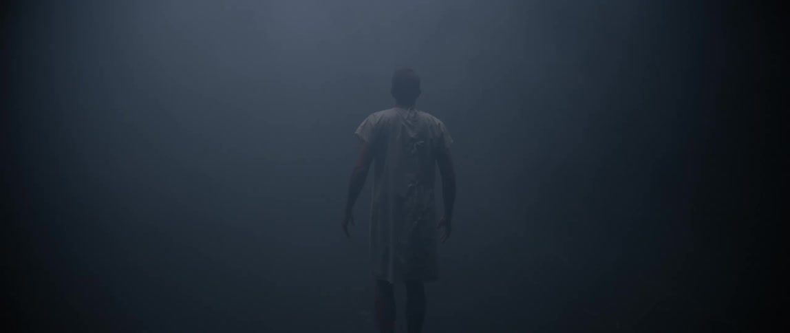 A person wearing a clinic patient gown is seen from behind walking through a dim misty environment