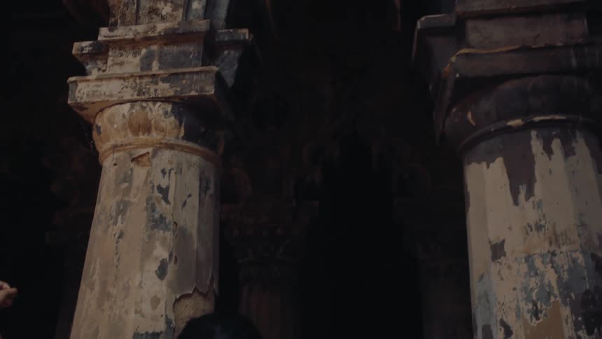 A woman standing in front of old building pillars with worn rough paint her left arm raised under the midday sun