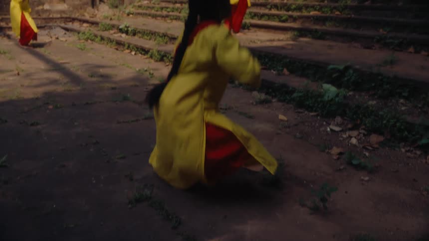 Three young women in yellow and red traditional attire perform a synchronized dance kneeling with outstretched arms on weathered stone steps in an outdoor setting