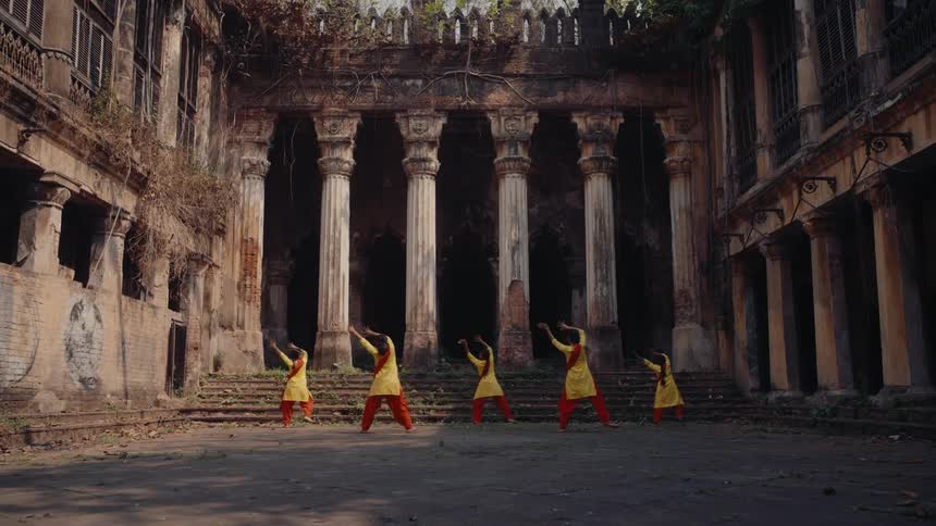 Five women in yellow and orange outfits are performing a synchronized dance in front of an old dilapidated building with tall columns and overgrown vegetation