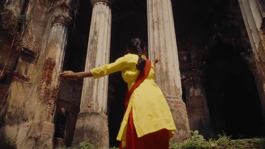 A woman in a traditional yellow and red outfit is walking up the steps of an old partially ruined building with large columns and overgrown vegetation