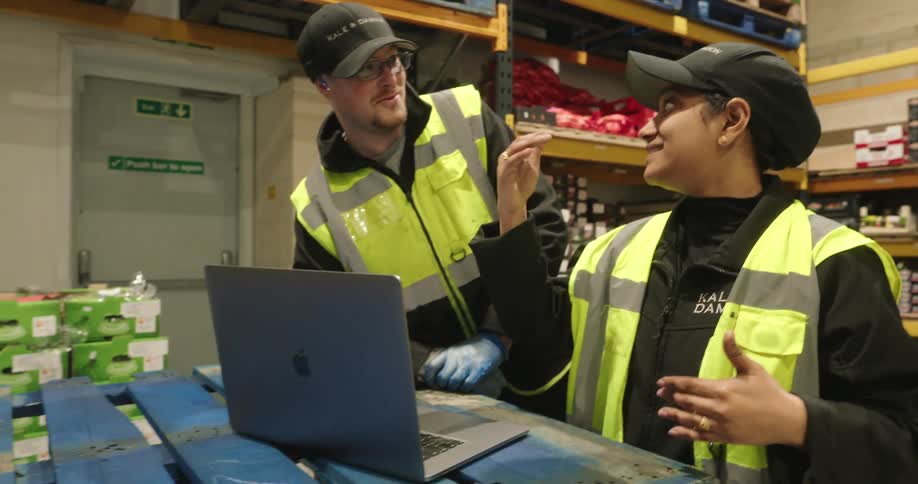Two warehouse workers in vests having a fun conversation together over a laptop