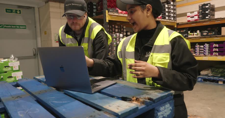 A man and woman stand inside a stockroom in front of a computer and have a conversation
