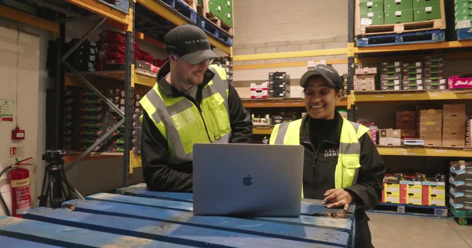 Two warehouse workers in safety vests laugh together while reviewing data on a laptop in a produce storage area