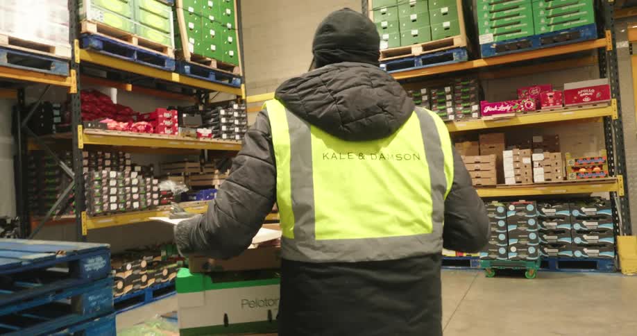 A warehouse worker in cold storage carries a box of yellow onions and places it on a cart while in a safety vest pushes a cart stacked with boxes of produce towards a set of large yellow doors likely leaving a cold storage area
