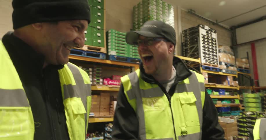 Three workerstwo men and one womanwearing green vests and hats are standing inside a warehouse engaged in a cheerful conversation and laughing together