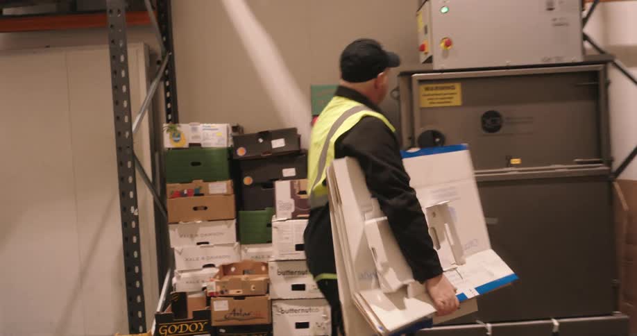 A group of four warehouse workers in safety vests handle paperwork and computer tasks in a storage area