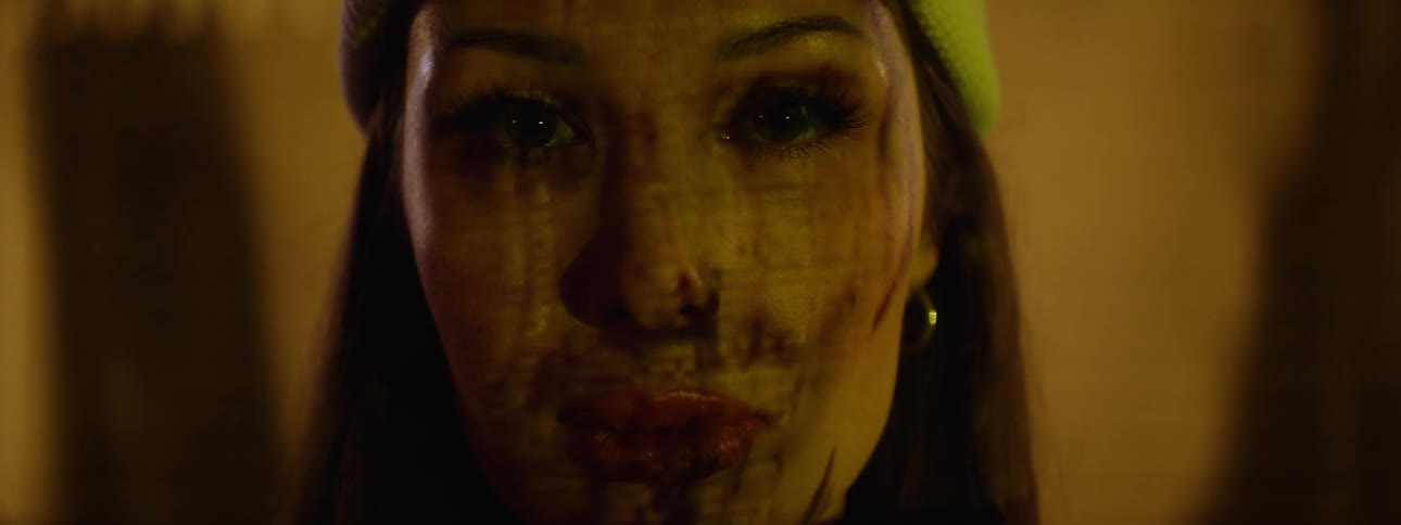 A woman looks directly at the camera her features overlaid by a projection of a mans face while behind her an led board displays light flashes and glitching giant faces