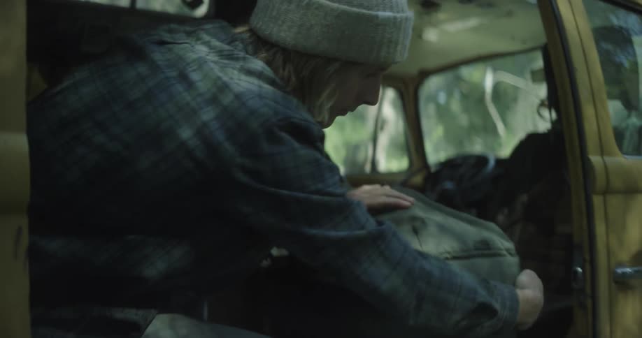 A young man inside a van with the door wide open is closing the zipper of a backpack at midday