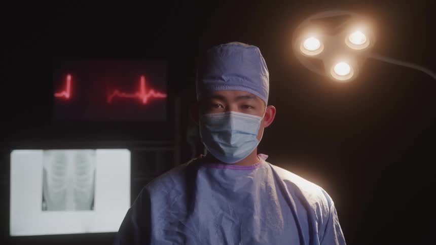An asian man dressed as a doctor wearing surgical scrubs and a mask standing in a dimly lit operating room with a heart monitor displaying a heartbeat and an xray of a chest in the background