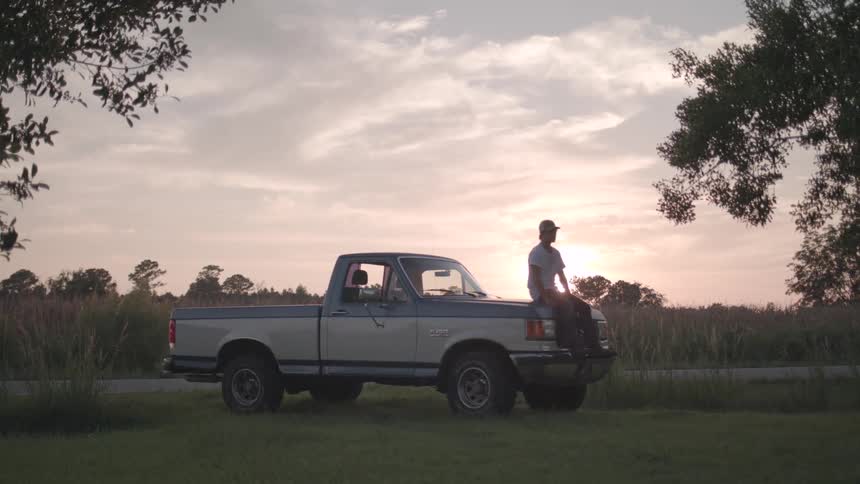 Man sits on the hood of his pickup truck across the highway from a crop field near sunset