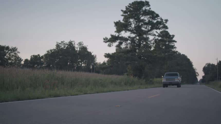 A beatup twotoned pickup truck drives on an asphalt road flanked by tall grass and trees under a pale sky