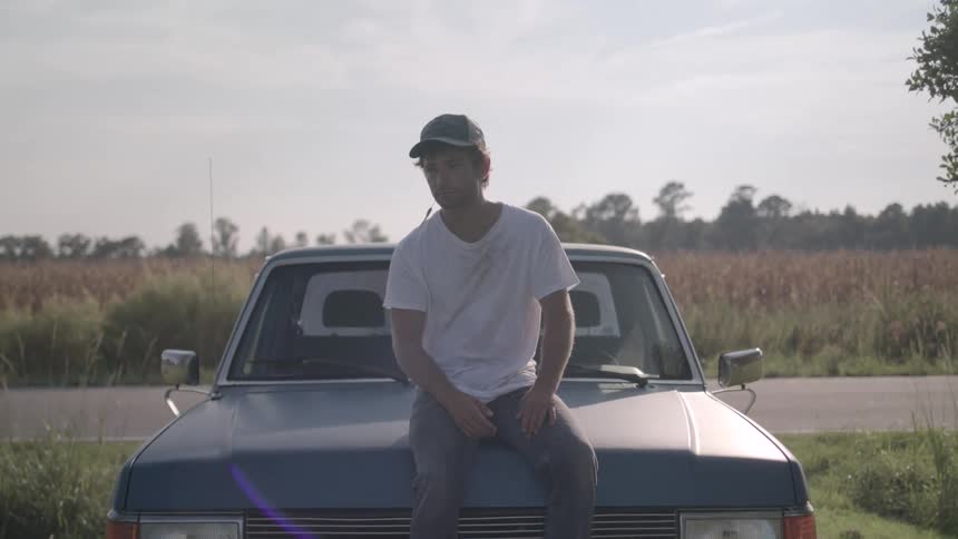 Man in cap and white shirt sitting in a parked car in a field