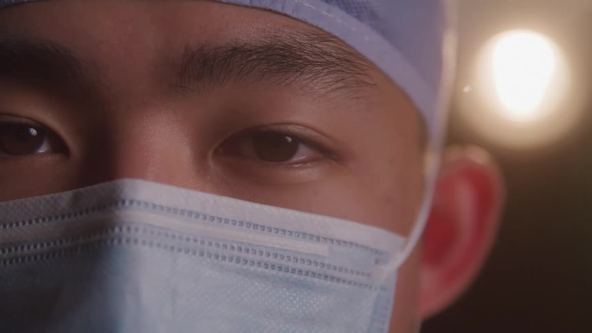 A doctor wearing a nose mask and surgical cap stands with a bright surgical light illuminating the area behind him