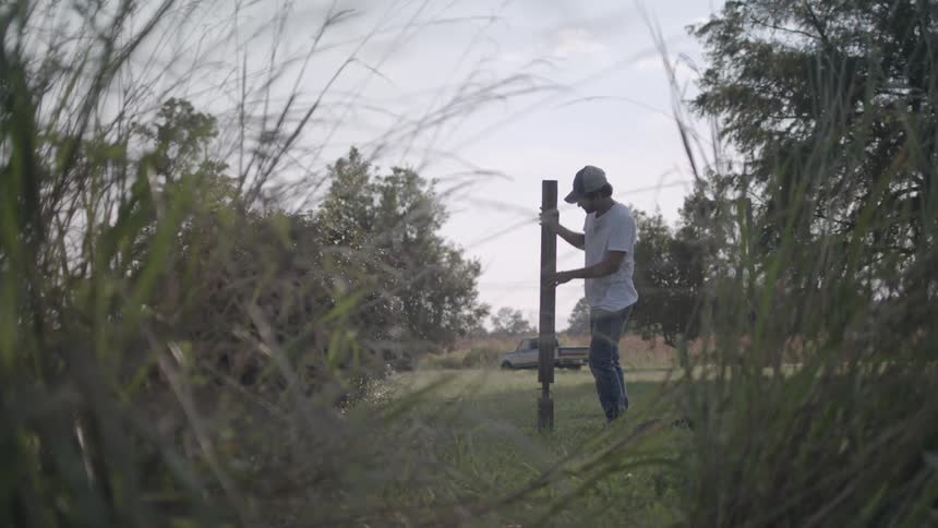 A man wearing a white tshirt and a hat is placing a piece of wood on the ground in a grassy field with a truck visible in the distance behind him at midday