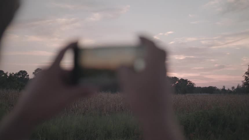 Pair of hands holding a smartphone and photographing a countryside scene in the late afternoon