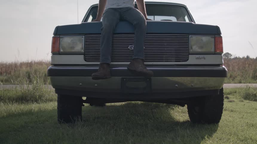A man wearing jeans and a white tshirt sits on the hood of a blue truck parked on a grassy field beside the road on a sunny day