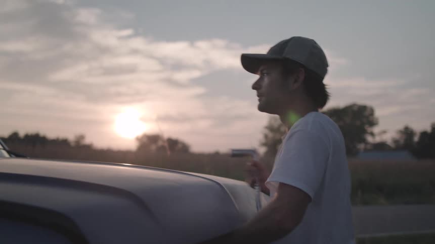 A man wearing a cap steps out of a pickup truck to open its hood silhouetted against a sunset backdrop