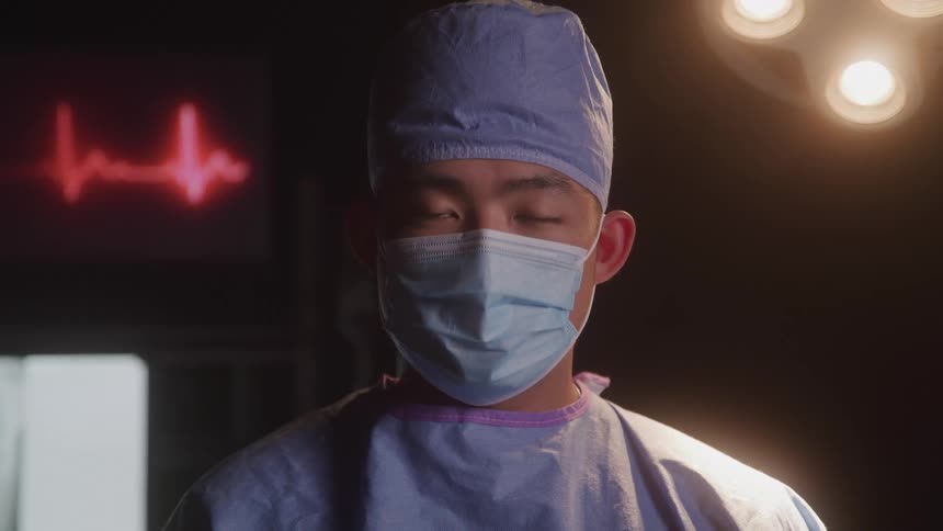 An asian man dressed as a doctor wearing surgical scrubs and a mask standing in a dimly lit operating room with a heart monitor displaying a heartbeat and an xray of a chest in the background