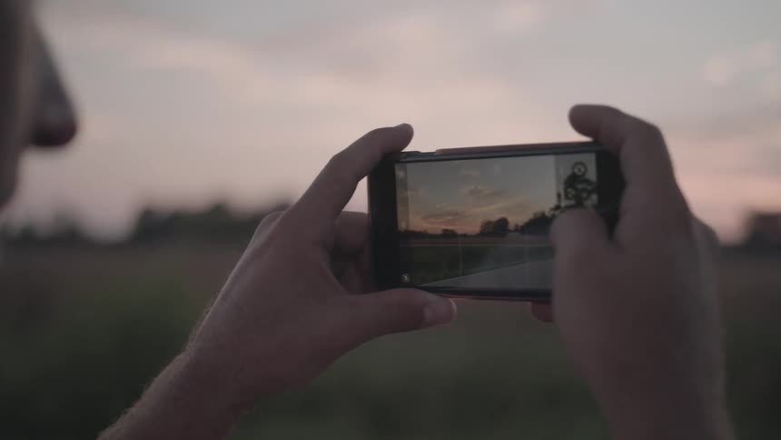 A person holds a smartphone horizontally using the screen to frame a photograph of a road stretching towards a horizon under a dusky sky