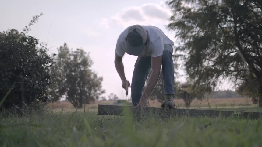 A man in a grassland holds a piece of wood down with one leg placed on top of it while cutting through it with a saw the area is near a road where a single car is seen driving by at midday