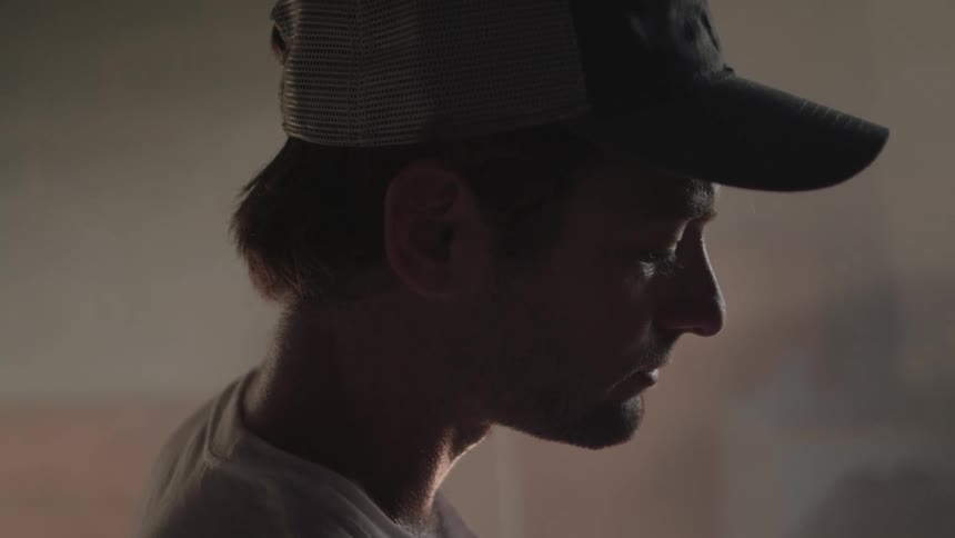 A closeup of a person wearing a baseball hat standing in a dimly lit room near a window in the morning
