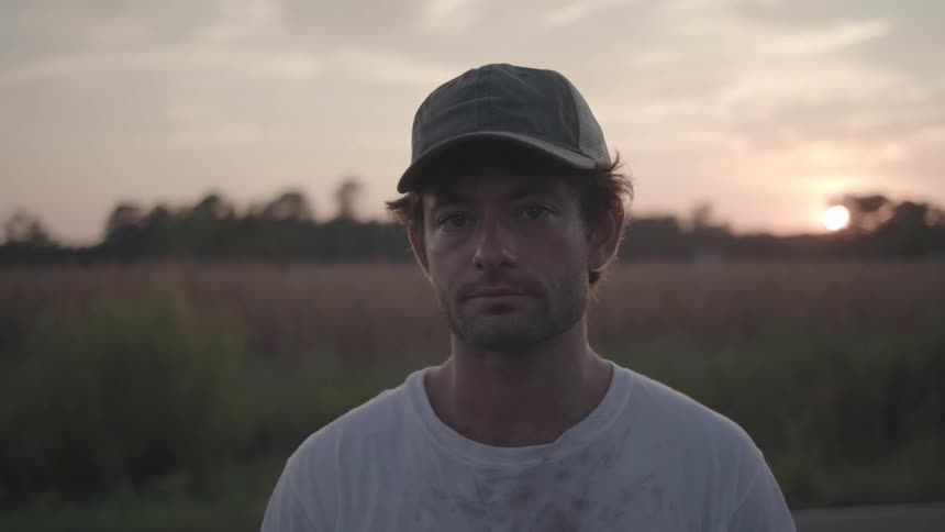 A man wearing a hat and a white tshirt with stains stands on a grassland at sunset