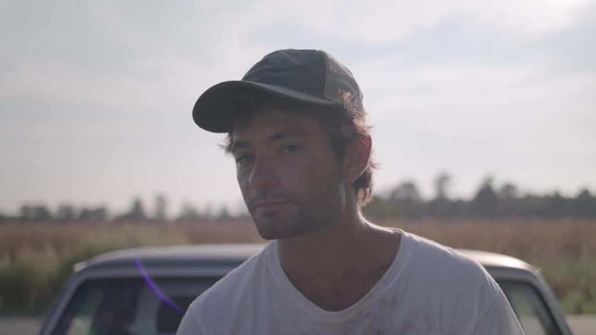 A person wearing a baseball cap and a white tshirt looks off to the left against a backdrop of a car and a hazy landscape