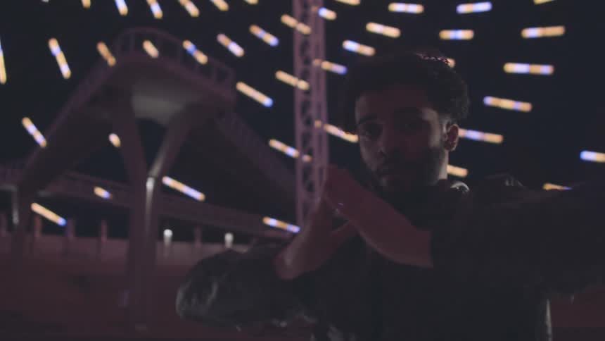 A person with curly dark hair and a patterned jacket dances with their hands near their head surrounded by blurred colorful lights in a dimly lit space