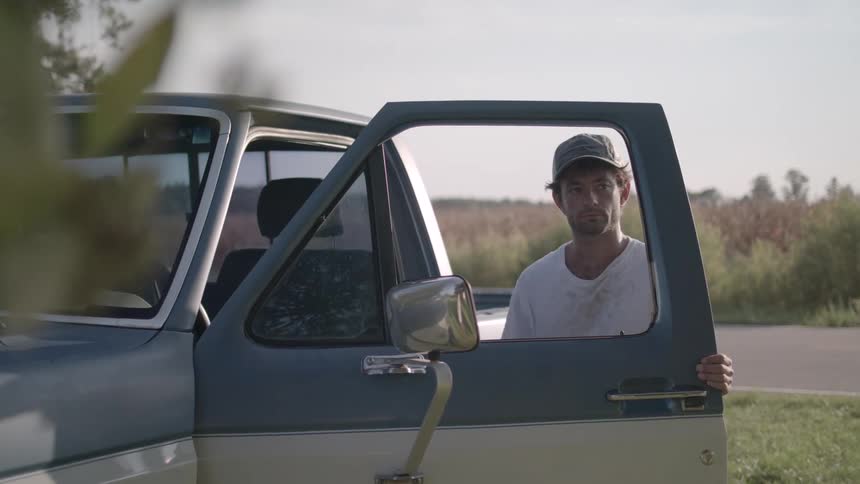 A man wearing a hat and a stained white tshirt opens the door of his truck pauses in deep thought then gets into the truck parked on a grassy field and shuts the door at midday