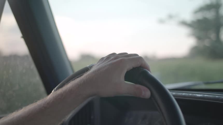 A mans arm holds the steering wheel while driving his vehicle on a road surrounded by vegetation and trees at midday