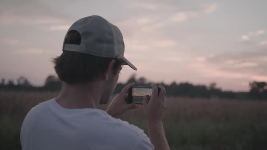 Man in cap holding smartphone sideways and photographing a field at dawn