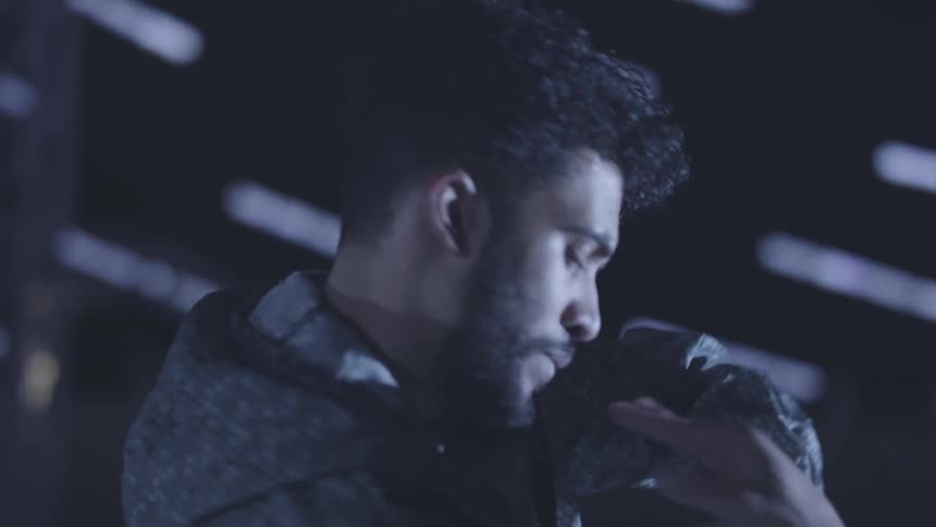 A dimly lit closeup shot captures a person with dark curly hair and a mustache moving their hands in fluid motions while dancing at night