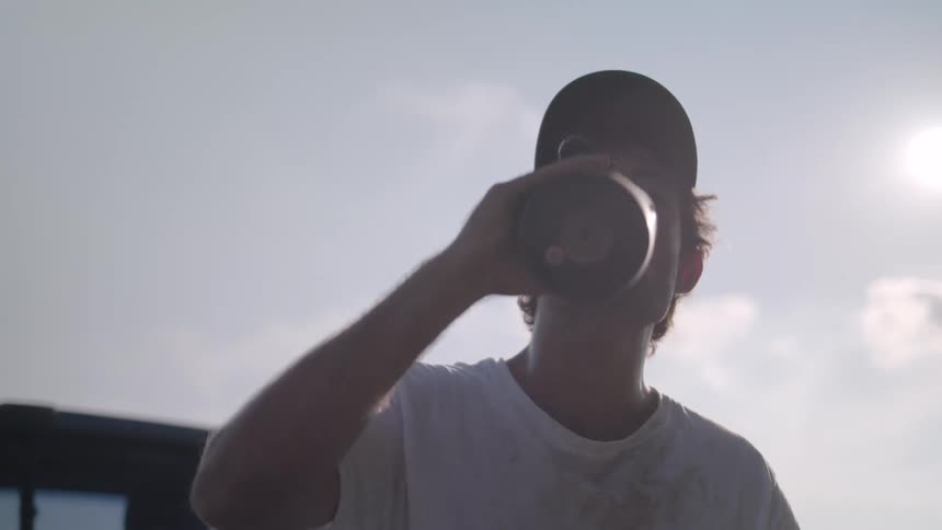 Man in cap outdoors rubbing his face and drinking liquid from a bottle