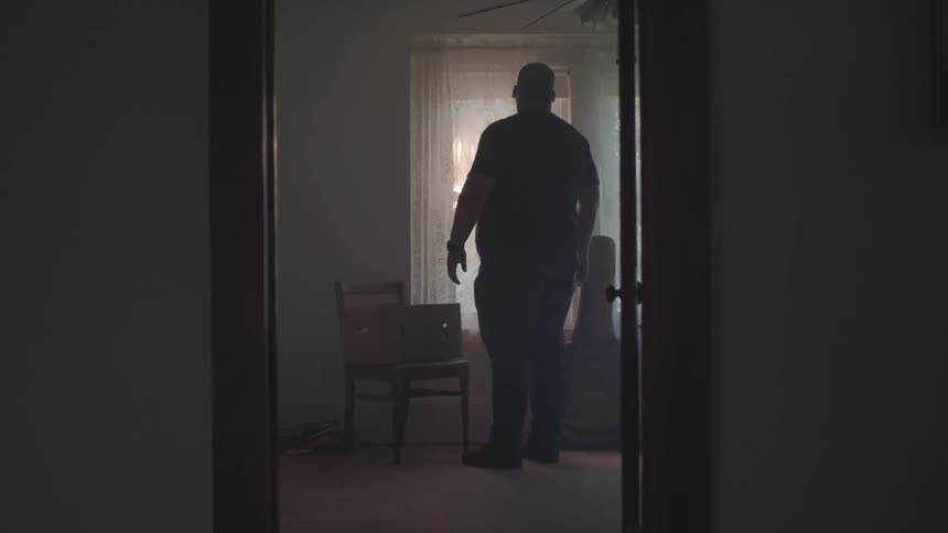 A man stands inside a room at midday looking through the window next to a chair