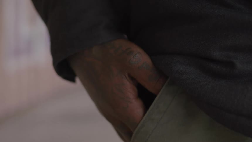 A tattooed hand is partially inserted into the pocket of lightcolored pants