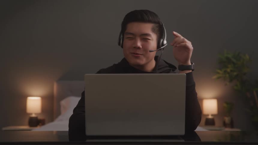 A man is in his bedroom wearing a headset on a conference call on his laptop talking and using hand gestures with two bedside lamps visible behind him
