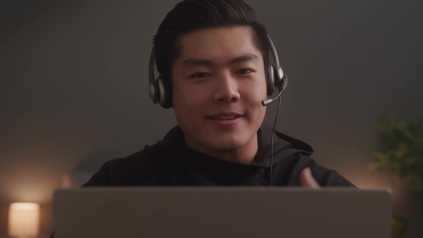 A young adult with dark hair wears a headset with a microphone looking at the camera over a laptop screen while gesturing animatedly as they explain something