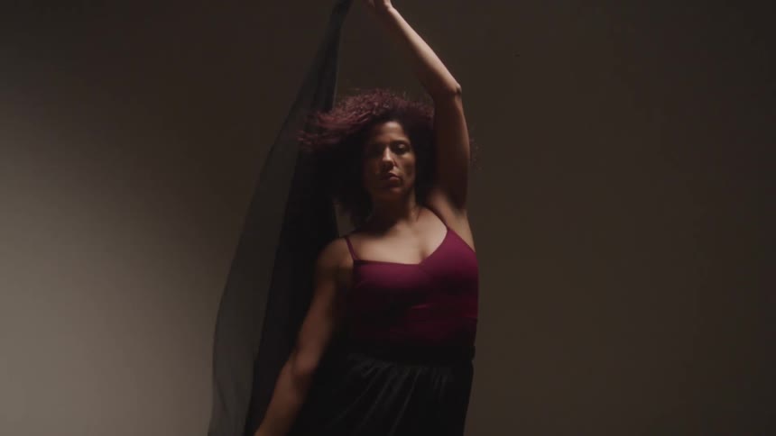 A woman with red curly hair poses for a photoshoot indoors holding a black light piece of cloth and wearing a red dress