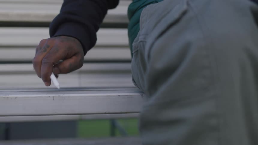 A tattooed hand belonging to a person dressed in gray pants grasps a piece of white chalk while resting on a bench