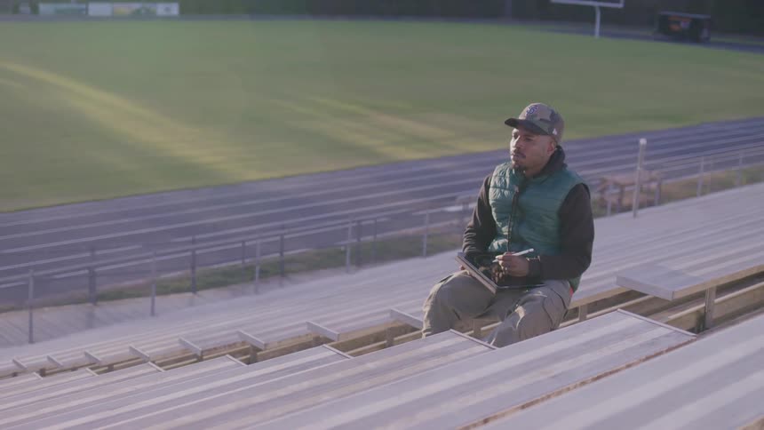 A black male sits on a bench in the seating area of a football field at midday writing in a notebook