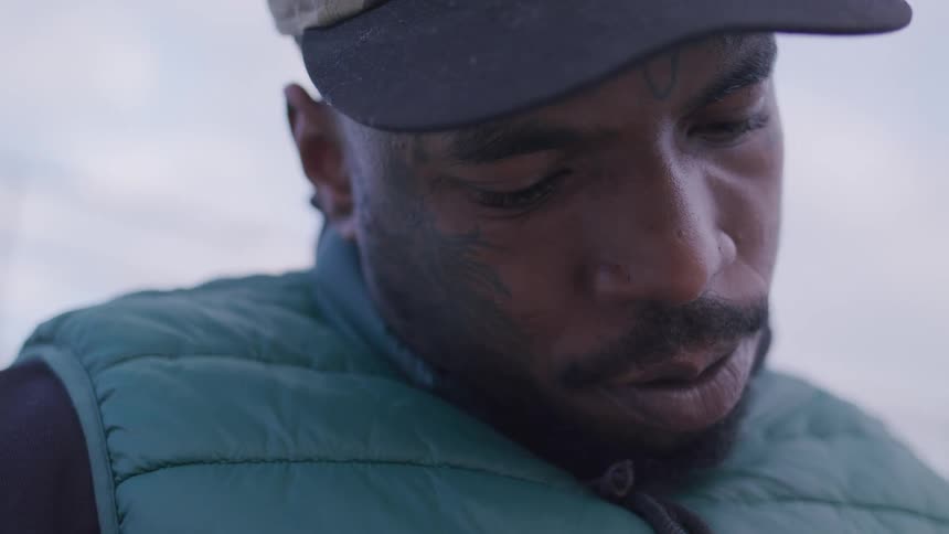A closeup shot of a man in a cap and padded vest looking down his expression focused and contemplative