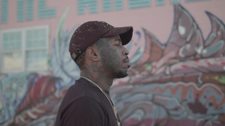 Tattooed man wearing a cap walking outdoors with a graffiti wall in the background