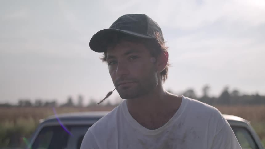 Man chews on a plant stem while sitting on his car near a field on an overcast summer day